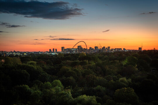 The illuminated arch of Wembley Stadium stands out against a vivid orange sunset sky above the London skyline, seen over a foreground of dense autumn treetops.