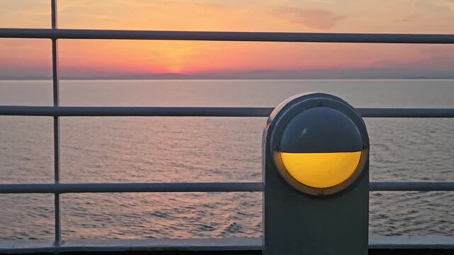 Sunset over calm sea, framed by cruise ship railings. Coin-operated binoculars glow warmly beside railing. Golden hues paint horizon, sky, and water. Ocean stretches wide. Stillness of scene, peaceful