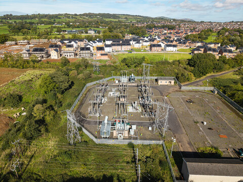 Aerial view of an electrical substation with metal transformers and power lines surrounded by green fields and a residential housing development in Lisburn, Northern Ireland, United Kingdom.