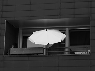 Black and white photograph focusing on a dark, modern building facade with a single balcony inset, featuring a prominent open white parasol and a window planter box. © Christian Bunge