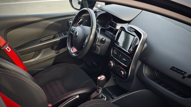 Interior view of a Renault Clio R.S. cockpit showing sport bucket seats with red seatbelts, leather steering wheel with paddle shifters, central infotainment screen, automatic gear selector 
