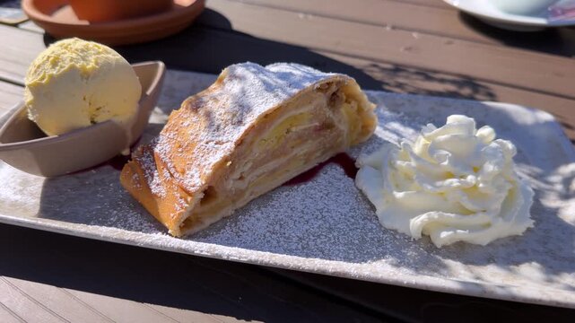 Traditional apple strudel dusted with powdered sugar and served with vanilla ice cream and whipped cream on a plate in warm sunlight.