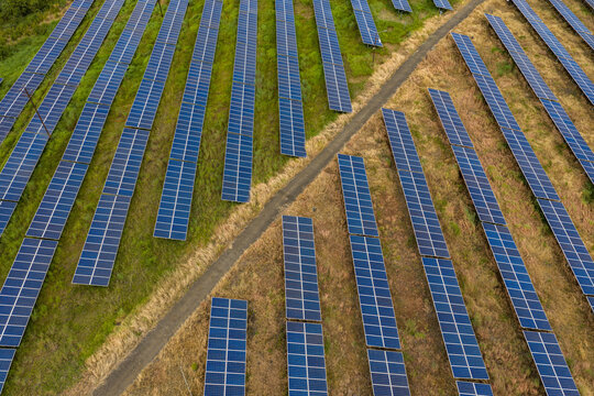 Aerial view of rows of blue solar panels arranged in a field with green grass and dry patches separated by a dirt path in Aundh, Maharashtra, India.