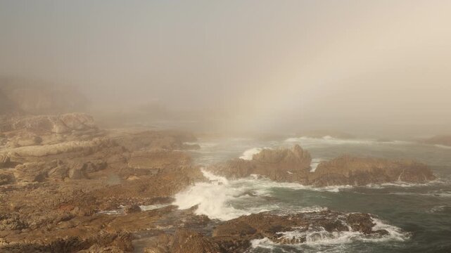 weather phenomenom coastal Hermanus, fog bank and rainbow as waves roll into rocky cliffs, South Africa