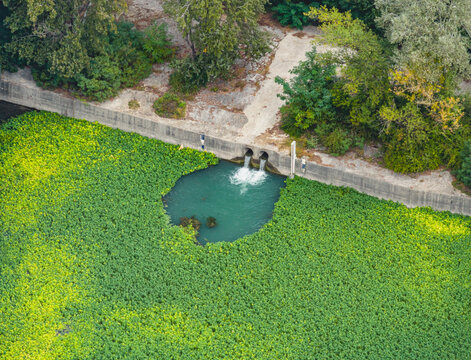 Aerial view of water discharge outlets flowing into a vegetation covered industrial drainage basin near a large manufacturing and processing facility in France.
