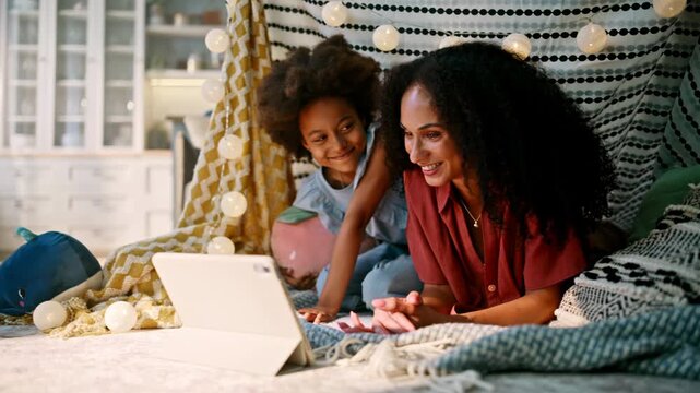Family blanket fort watching tablet movie. Mom daughter laughing together night.