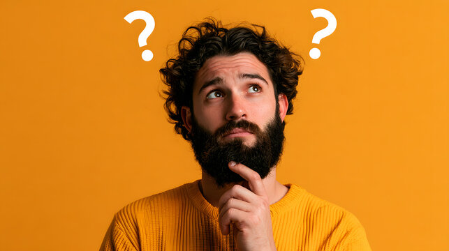 Young bearded man looking up with a confused facial expression, touching his beard in doubt, thinking about a problem, isolated on a bright orange background with floating white question mark symbols