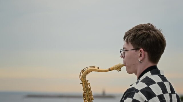 Caucasian young adult man playing saxophone outdoors against coastal skyline. Clip suits music education commercials, youth lifestyle campaigns, and urban culture promo videos