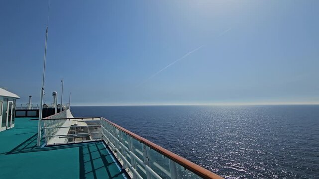 Cruiser deck faces open ocean under clear sky. Sunlight glints across calm waters. Deck railing frames horizon. Sky shows distant airplane trail. Cruise ship moves slowly on sea