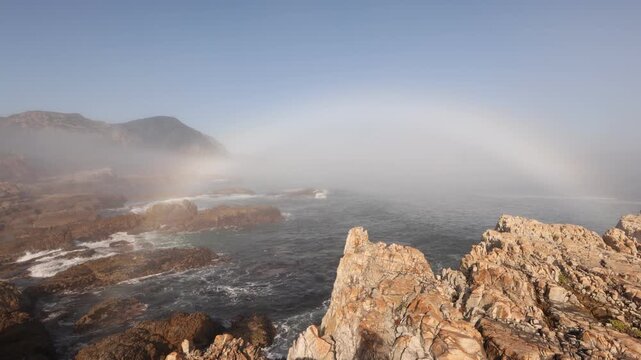 weather phenomenon: coastal Hermanus, fog bank and rainbow as waves roll into rocky cliffs, South Africa