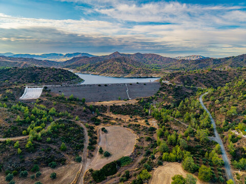 Aerial view of Dipotamos Reservoir showing the dam wall, rugged hills, sparse vegetation, and a winding road in Dimos Lefkaron, Larnaca, Cyprus.