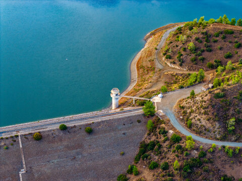 Aerial view of Dipotamos Reservoir with its distinctive water intake tower, concrete dam wall, and surrounding arid hills with sparse vegetation in Dimos Lefkaron, Larnaca, Cyprus.