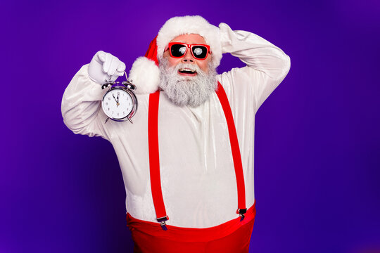 Santa Claus holding an alarm clock for Christmas countdown, smiling in red suspenders and sunglasses against a purple background