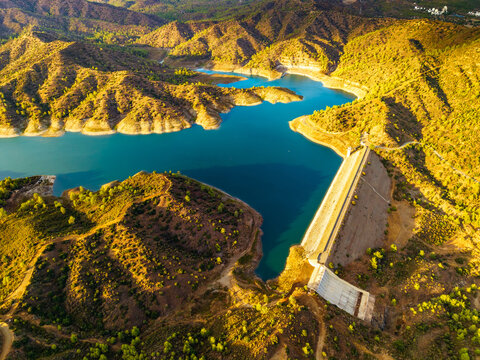 Aerial view of Dipotamos Reservoir with its large dam structure and turquoise water surrounded by golden sunlit hills and Mediterranean vegetation in Dimos Lefkaron, Larnaca, Cyprus.