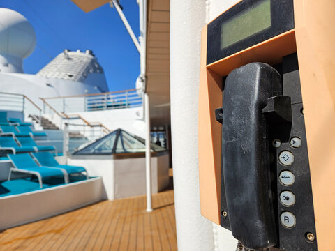 Cruise ship deck with phone and blue seating. Wooden floor, clear sky, modern design. Phone mounted on white wall, digital display. Glass roof and stairs visible in background