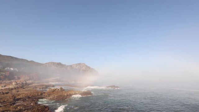 weather phenomenon: coastal Hermanus, fog bank and rainbow as waves roll into rocky cliffs, South Africa