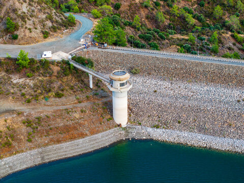 Aerial view of the Dipotamos Reservoir featuring its concrete dam tower, surrounding rocky hills with pine trees, and a winding road in Dimos Lefkaron, Larnaca, Cyprus.