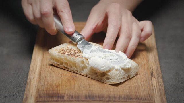 chef spreads fresh Italian ricotta cheese on slice of toasted ciabatta bread on wooden kitchen board set on black tabletop. The process of preparing Italian tomato and ricotta bread; food closeup