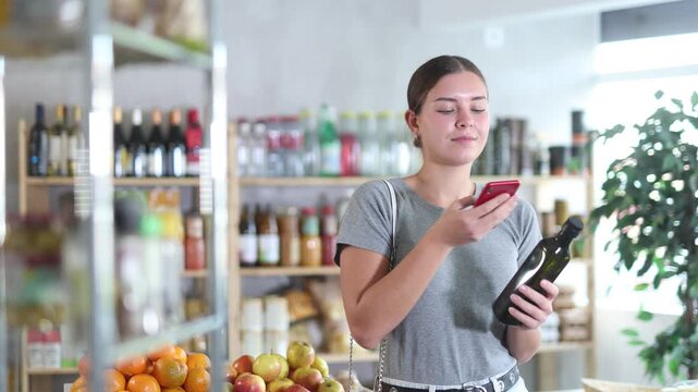 Girl in store scan QR code and study oil assortment. With help of mobile app, buyer receive information about olive oil product. She check manufacturer data in registry database of national agency 