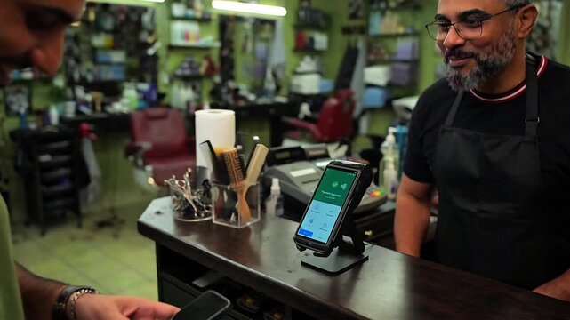 Customer making a contactless payment with a mobile phone at a barbershop checkout counter. Panning video footage of a successful digital transaction with a friendly male barber