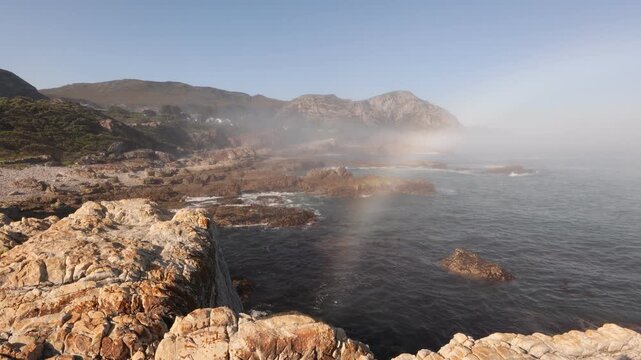 weather phenomenon: coastal Hermanus, fog bank and rainbow as waves roll into rocky cliffs, South Africa