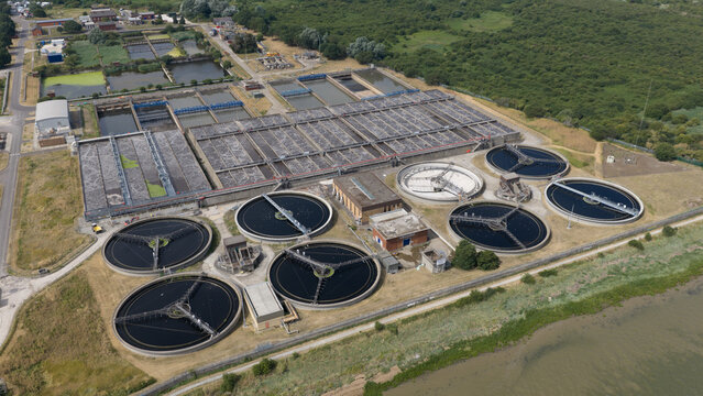 Aerial view of a large wastewater treatment plant featuring circular sedimentation tanks and rectangular basins surrounded by green landscape in Dartford, England, United Kingdom.