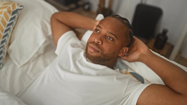 Man reclining on bed wearing white t shirt with hands behind head and gaze upward in bedroom building; calm reflection.