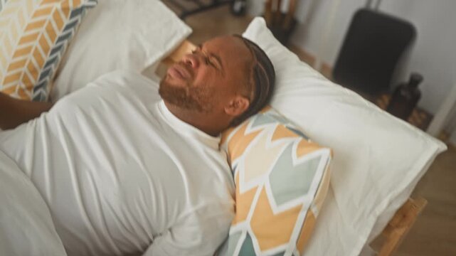Man on bed holds neck, wincing and rubbing nape while leaning on patterned pillow in a building; discomfort morning tension.
