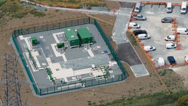Aerial view of Thurrock Flexible Generation Substation with green equipment containers in a fenced compound and an adjacent parking area with white vans in Tilbury, England, United Kingdom.
