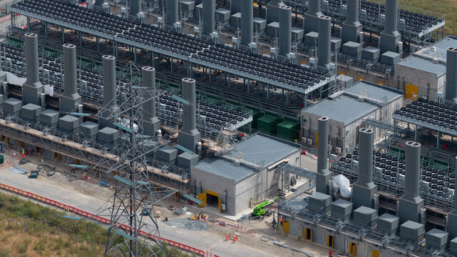 Aerial view of the Thurrock Flexible Generation Substation featuring rows of industrial cooling fans, tall exhaust stacks, and an electrical transmission tower in Tilbury, England, United Kingdom.