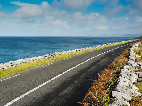 Asphalt road curves beside endless ocean. Grass grows between stones and pavement. Sky holds soft clouds above the sea. Road leads toward distant horizon. Perfect for travel, nature, or coastal themes