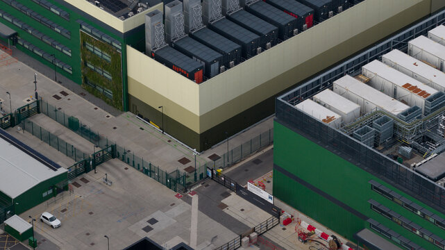 Aerial view of the NTT LON1B Data Centre featuring industrial buildings with green facades, rooftop cooling units, and a secure fenced yard in Dagenham, England, United Kingdom.