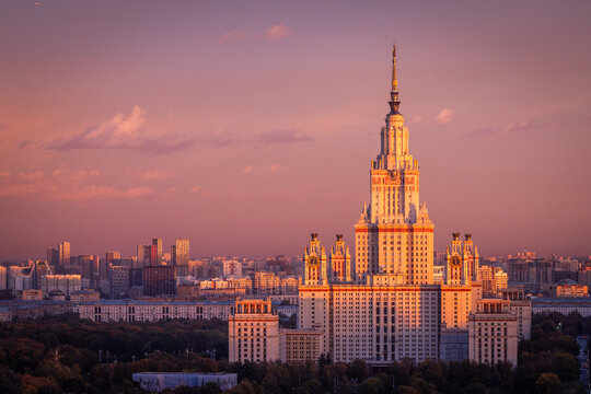 Aerial view of Moscow State University, an iconic Stalinist skyscraper illuminated by warm golden hour light, surrounded by lush parkland and the city skyline of Moskva, Moskva, Russia.