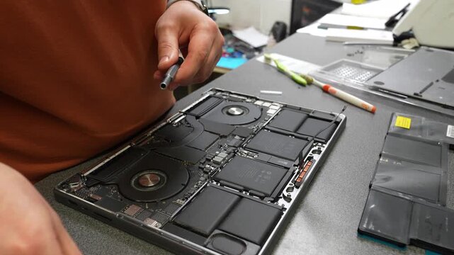 Technician uses a screwdriver to repair an opened laptop, exposing cooling fans, battery cells, and internal components on an electronics workbench
