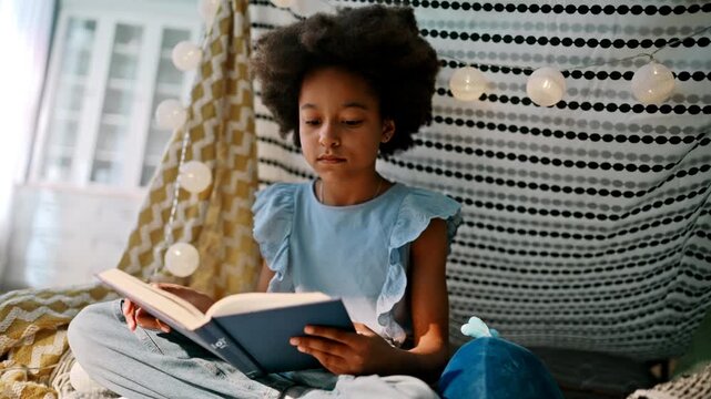 Mixed race girl reading book cozy home tent. Smiling kid enjoying fairy tale.