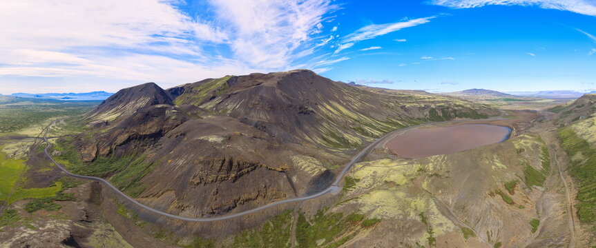 Aerial view of Almannagja rift valley featuring a winding road through rugged volcanic terrain, moss-covered lava fields, and a small lake in Blaskogabyggd, Iceland.