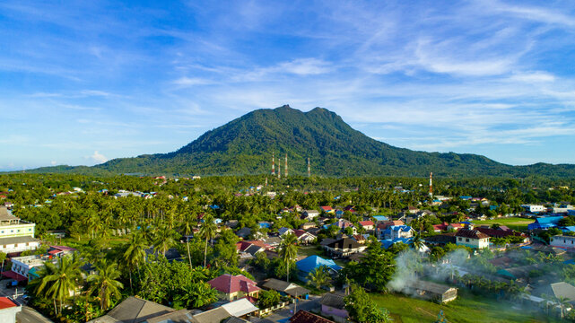 Aerial view of a town nestled at the base of a lush green mountain in Indonesia, featuring palm trees, residential houses, and communication towers under a blue sky.