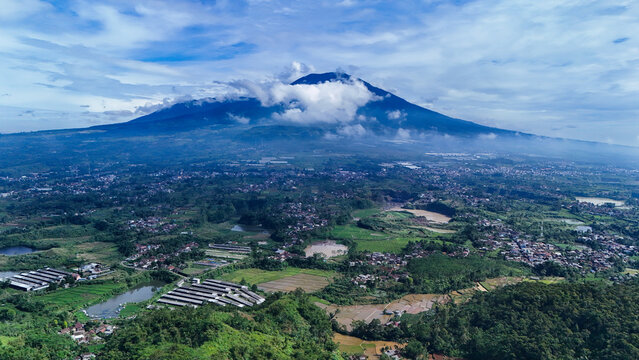 Aerial view of Mount Gede Pangrango National Park with lush green rice fields, rural settlements, and a sand mining site under a cloudy blue sky in Sukalarang, West Java, Indonesia.