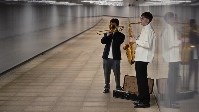 Young Caucasian musicians busking with saxophone and trombone in pedestrian underpass. Clip perfect for urban lifestyle ads, music festival promotion, and arts documentaries