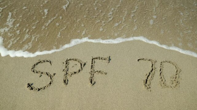 Spf 70 sun protection factor writing washing away. Gentle ocean wave washing away the text spf 70 handwritten on the wet sand of a beach, symbolizing the need to reapply sunscreen for effective sun