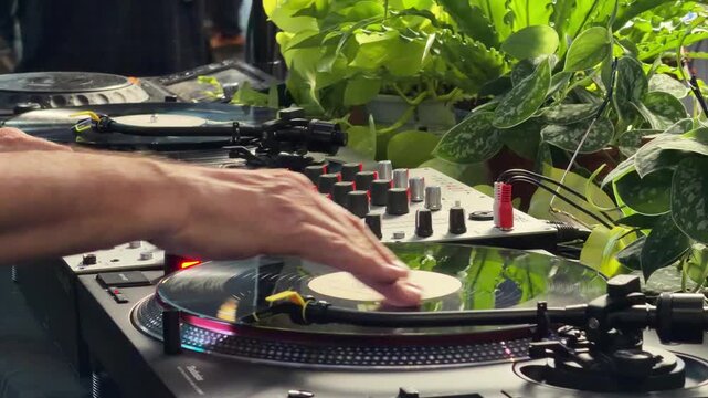Closeup of DJ hands mixing music on a vinyl turntable and sound mixer surrounded by lush green plants, party, nightlife and electronic music event concept