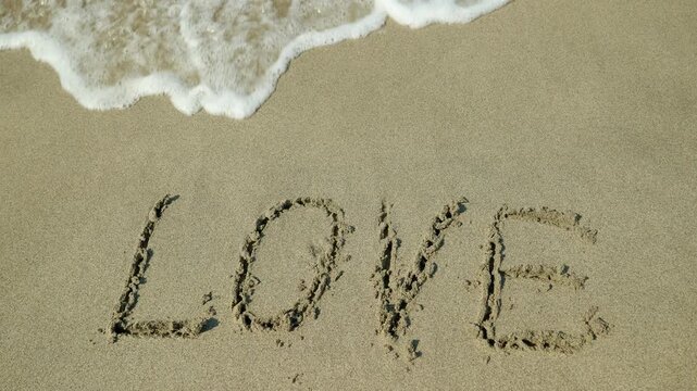 Ocean wave washing away the word love from the sand. Gentle sea foam from an ocean wave slowly washing away the word love handwritten on a sandy beach, symbolizing themes of heartbreak, impermanence