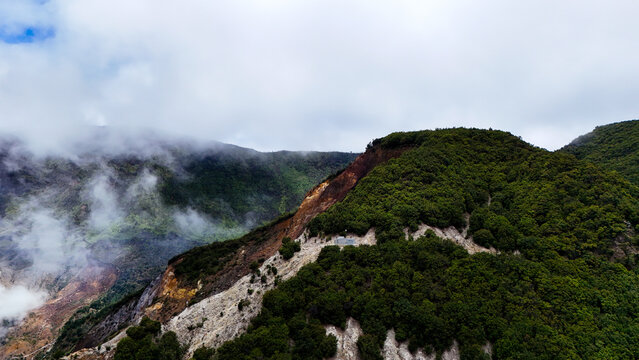 Aerial view of Mount Papandayan featuring volcanic slopes with sulfurous deposits, dense green forest, and drifting clouds over the rugged landscape in Garut, West Java, Indonesia.