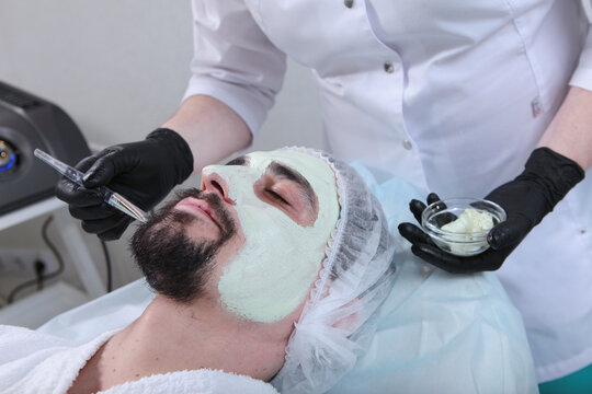 Man with greenish mask on his face in cosmetology office. Male client with beard receives greenish face mask. Men's cosmetology. Specialist in white uniform holds brush and glass bowl