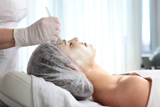 Applying cream mask to client's face in salon. Professional applies creamy mask on client face using brush. Gloved hand holds glass bowl. Bright clinical setting. Client relaxed on treatment bed