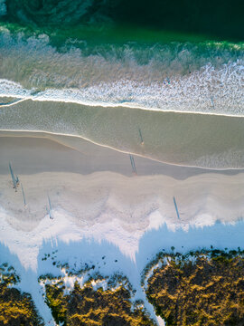 Aerial view of Middleton Beach with turquoise ocean waves washing onto the white sand, long shadows of people walking, and coastal vegetation in Albany, WA, Australia.