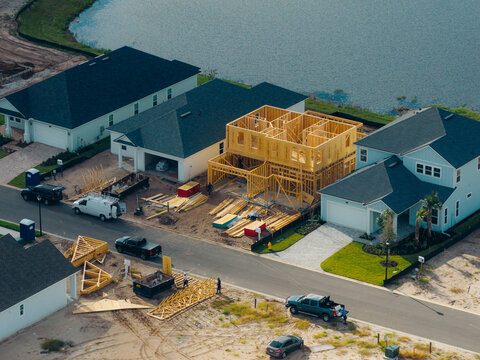 Aerial view of a residential construction site with wooden framing of a new house, completed suburban homes, a paved street, and a nearby lake in Jacksonville, Florida, United States.