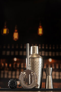 Shaker jigger strainer muddler on black mat. Blurred bar background with bottles and warm light. Dark background. Copy space
