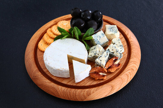Cheese board with brie, blue cheese cubes, crackers, black olives, walnuts, mint on round wooden platter. Dark background