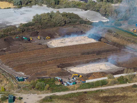 Aerial view of a composting facility with large piles of organic waste, heavy machinery, and smoke rising from the site in the United Kingdom.